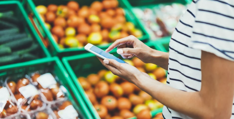 Persona usando un móvil en un supermercado, buscando consejos para ahorrar en la cesta de la compra.