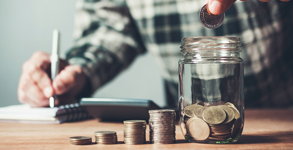 Mano depositando monedas en un frasco mientras toma notas, mostrando cómo ahorrar hasta 3.000 euros al año.