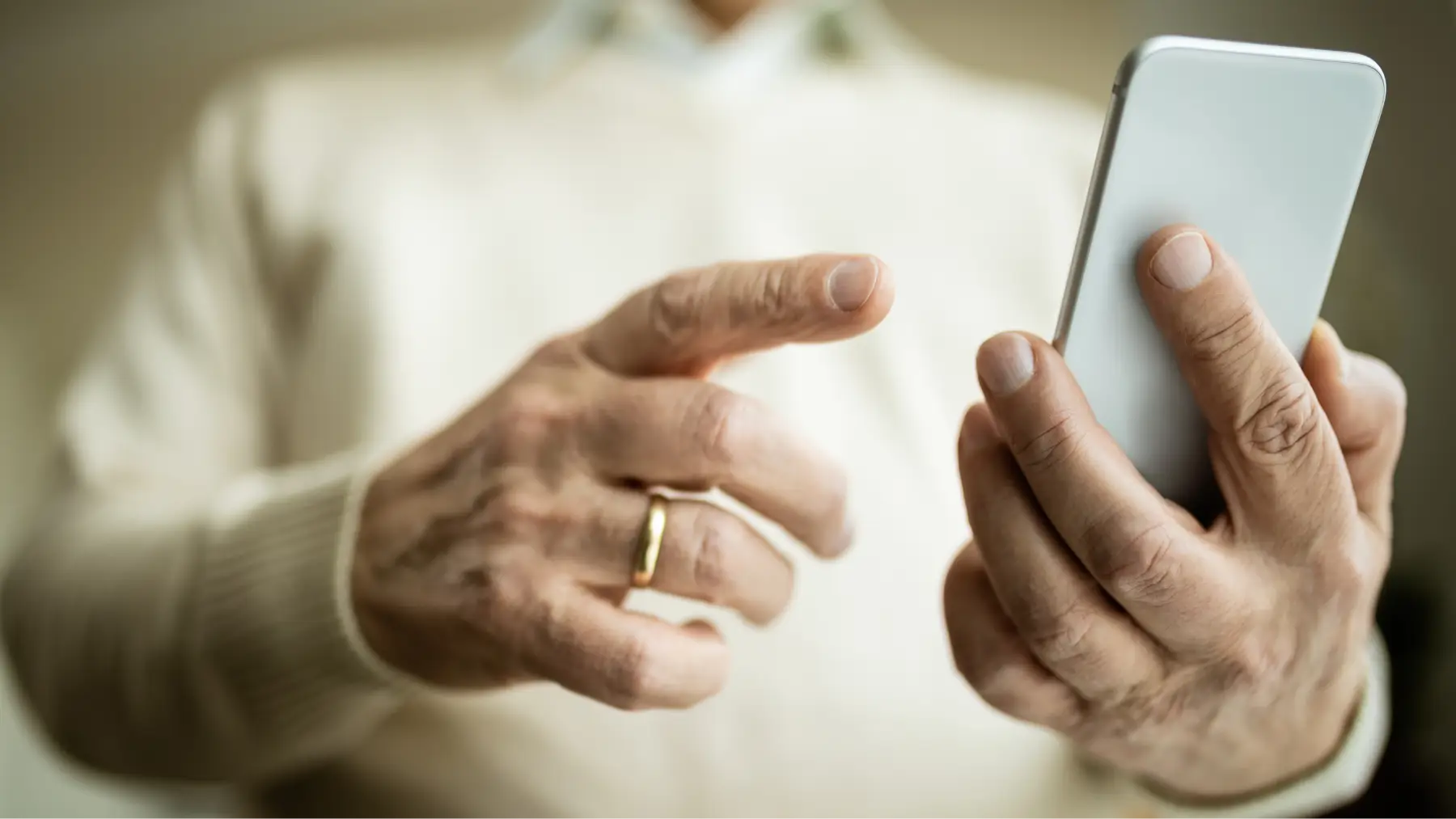 Primer plano de las manos de una persona mayor sosteniendo y utilizando un teléfono inteligente blanco.