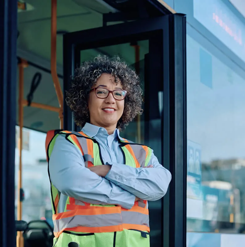 Conductora de autobús sonriendo con chaleco reflectante junto al vehículo.