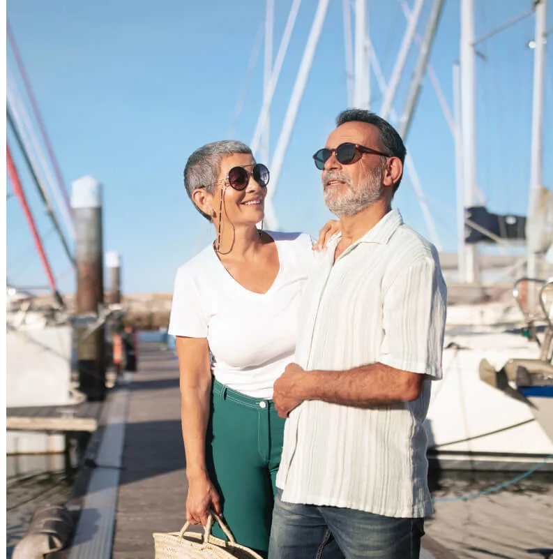 Pareja adulta sonriente paseando por un puerto deportivo, rodeada de barcos y veleros en un día soleado.