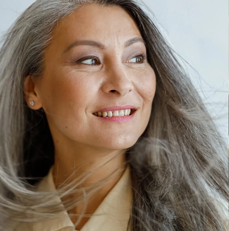 Un primer plano de perfil de una mujer mayor con el pelo largo y plateado, que sonríe de forma optimista mientras mira hacia la derecha contra un fondo blanco minimalista.