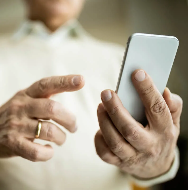 Primer plano de las manos de una persona mayor sosteniendo y utilizando un teléfono inteligente blanco.