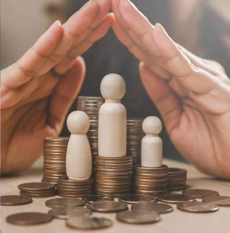 Manos protegiendo figuras de madera sobre pilas de monedas.