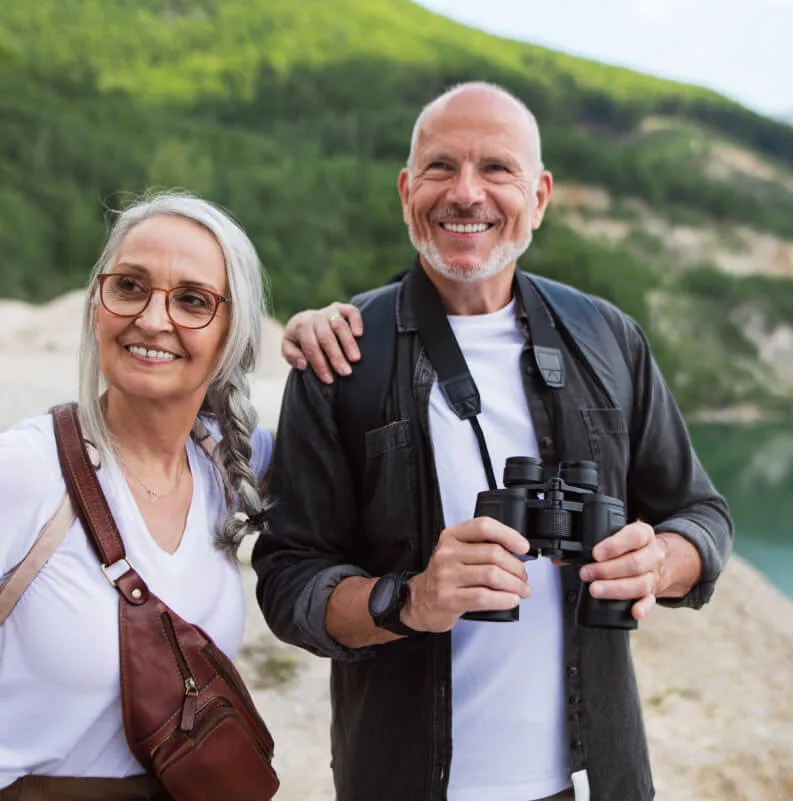 Pareja de jubilados sonriendo y mirando a la distancia durante una excursión en la montaña con lago al fondo