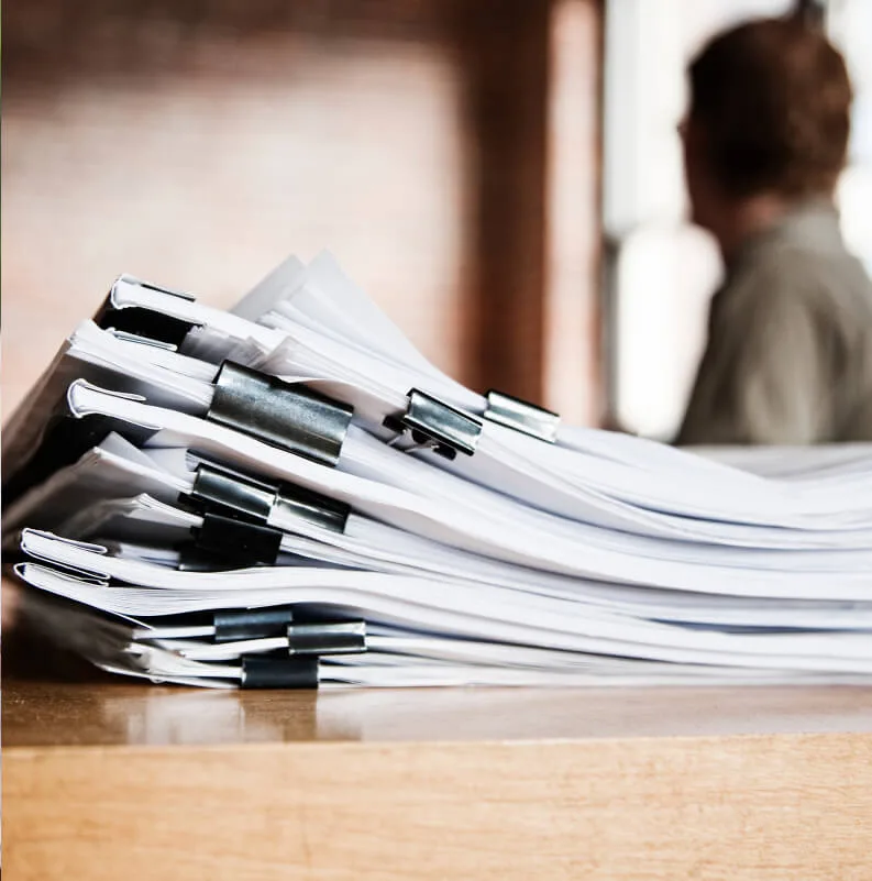 Primer pla d'una gran pila de documents de paper blancs sobre una taula de fusta, subjectes per múltiples pinces subjectapapers negres. Al fons, borrosa, es veu la silueta d'una persona d'esquena i una paret de maó.