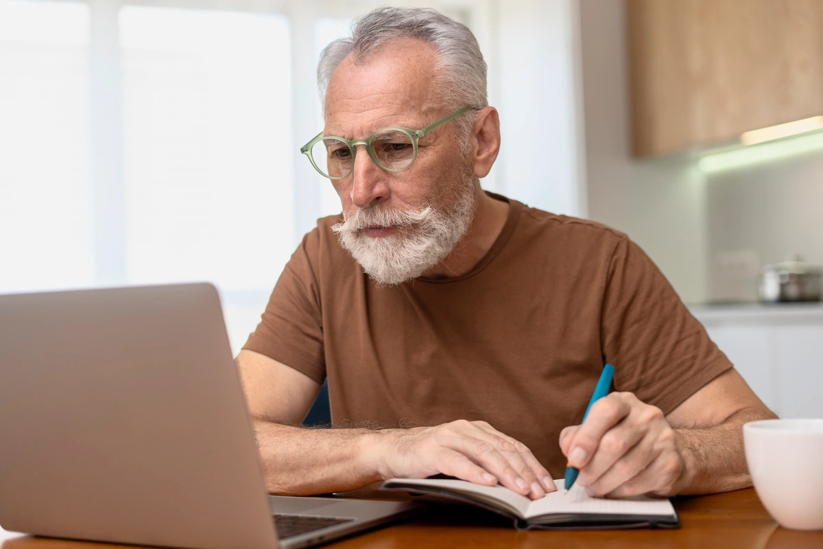 Hombre estudiando a los 50 frente al portátil tomando apuntes en una libreta.