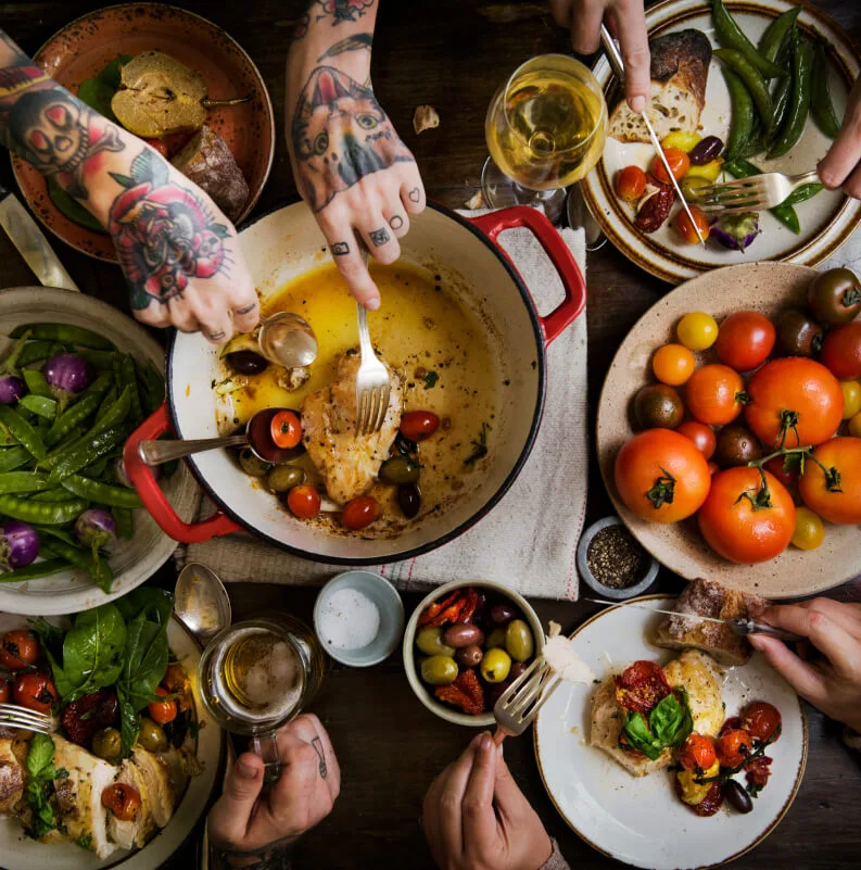Vista cenital de varias personas compartiendo una comida saludable con alimentos propios de la dieta mediterránea, como tomates, verduras y aceite de oliva.
