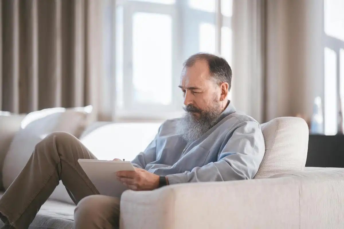 Home amb barba assegut en un sofà escrivint concentrat en una llibreta.