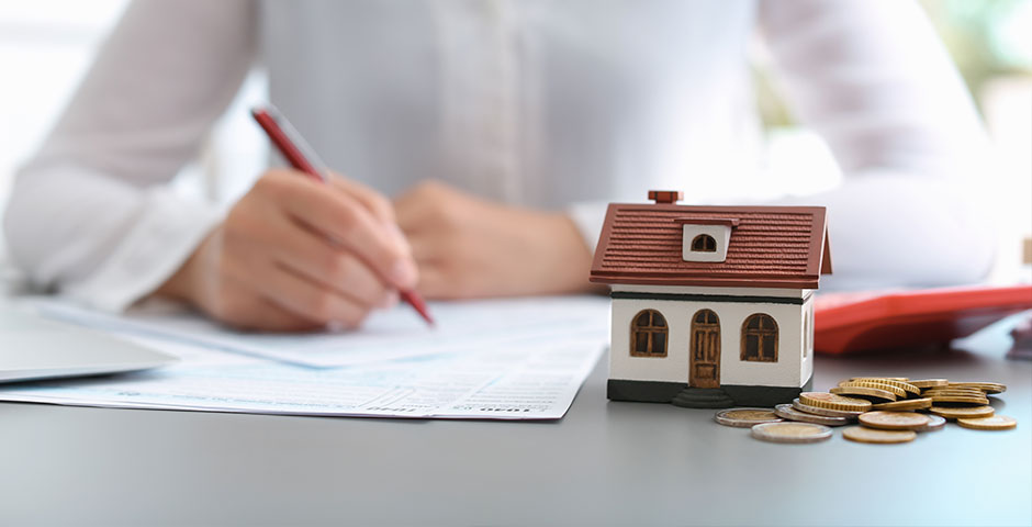 Persona completando documentos fiscales junto a una casa en miniatura y monedas.