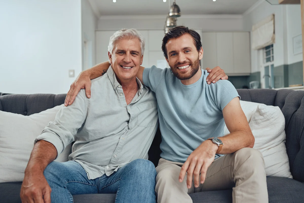 Padre e hijo sonriendo juntos en un sofá, hablando sobre el impuesto de sucesiones y donaciones.
