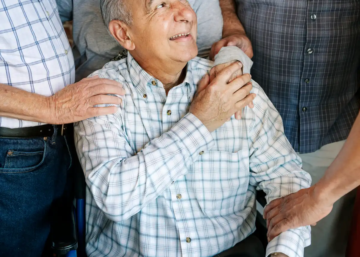 Hombre mayor en silla de ruedas sonriendo acompañado de personas que le muestran apoyo y respeto.