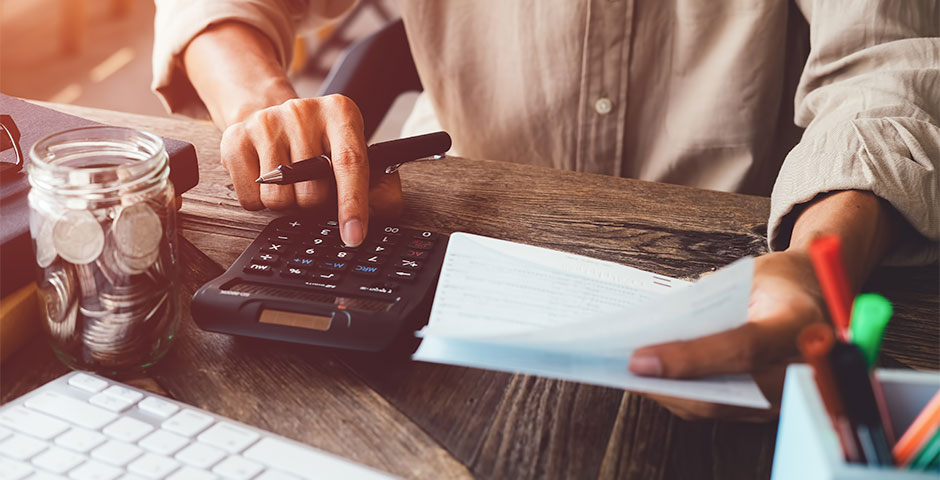 Persona usando calculadora mientras revisa documentos financieros en una mesa con frasco de monedas.