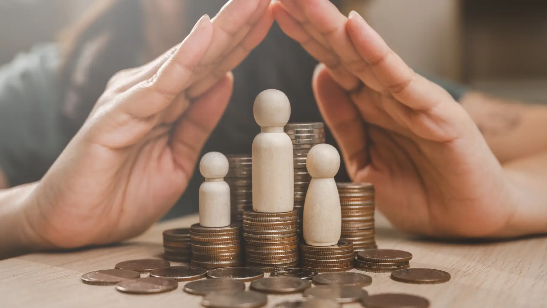 Manos protegiendo figuras de madera sobre pilas de monedas.