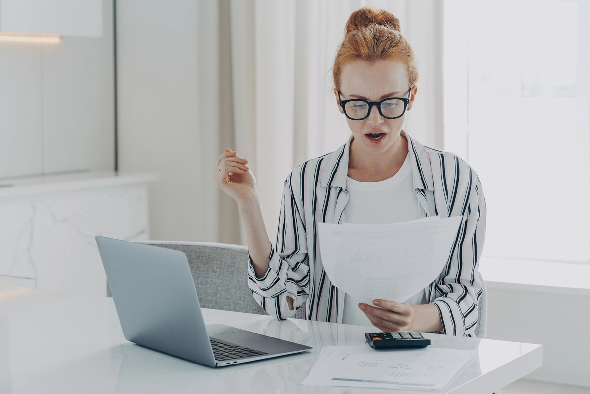 Mujer joven con gafas sorprendida revisando papeles sobre su plan de ahorro en el escritorio.