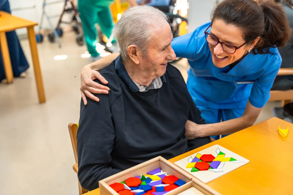 Home gran amb símptomes d'Alzheimer en teràpia cognitiva acompanyat de la cuidadora.