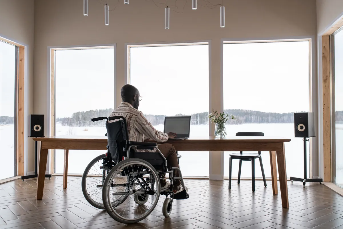 Persona en silla de ruedas en el salón de casa usando el ordenador frente a unos ventanales con incapacidad permanente absoluta.