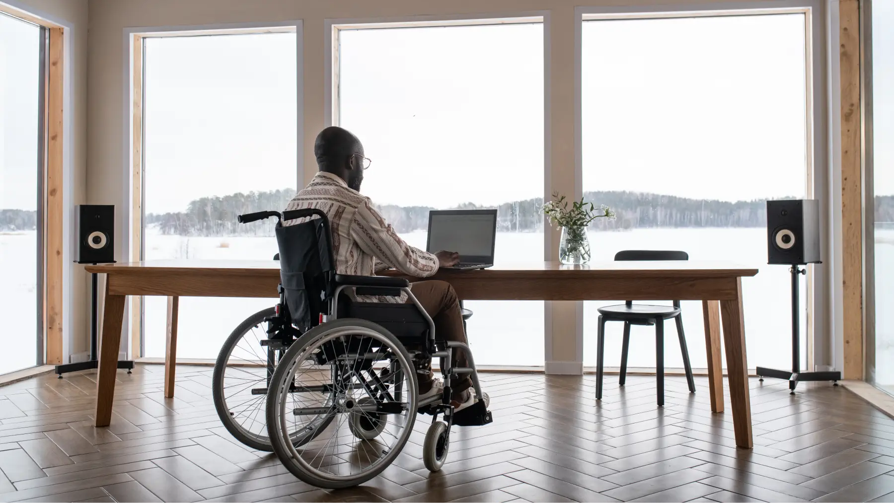 Persona en silla de ruedas en el salón de casa usando el ordenador frente a unos ventanales con incapacidad permanente absoluta.